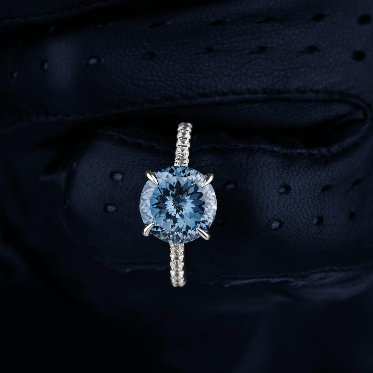 Close-up of a diamond engagement ring with a round center stone and a pavé diamond band on a black textured background.