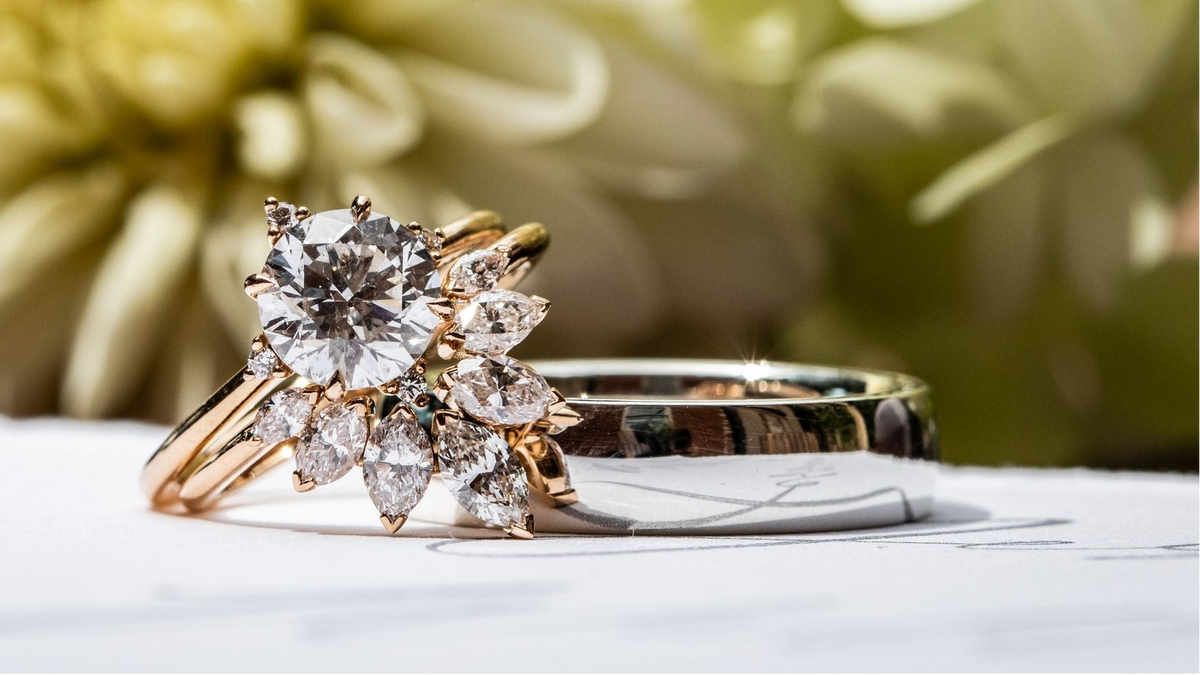 A close-up of a gold ring with a large round diamond and smaller marquise-cut diamonds, alongside a plain gold band, resting on a white surface with a blurred yellow flower in the background.