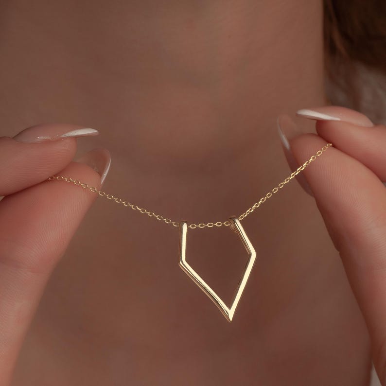 A close-up of a person holding a delicate gold chain necklace with a geometric open-frame pendant against their chest.