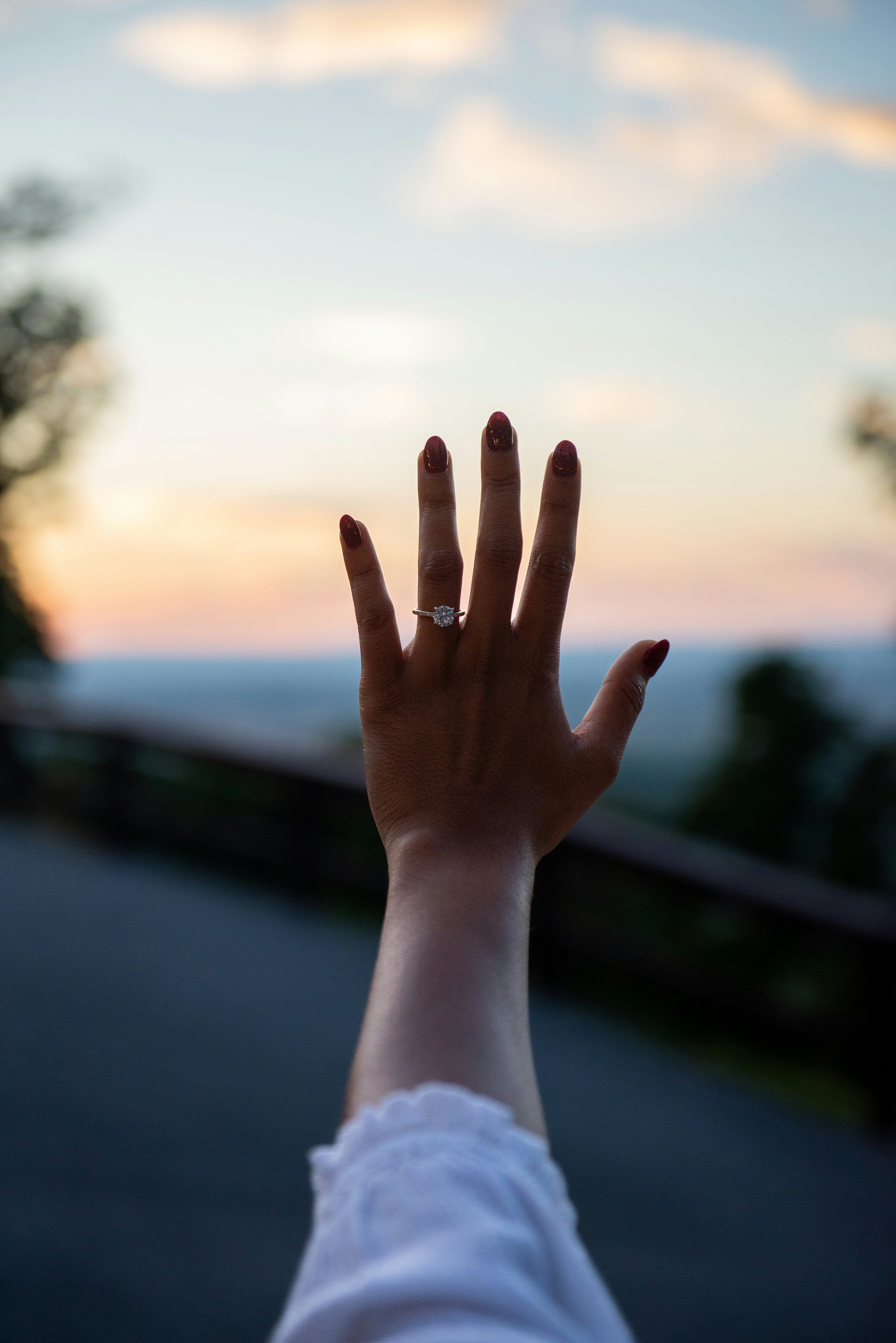 Hand raised against a blurred sunset background showcasing a bespoke diamond engagement ring on the ring finger.