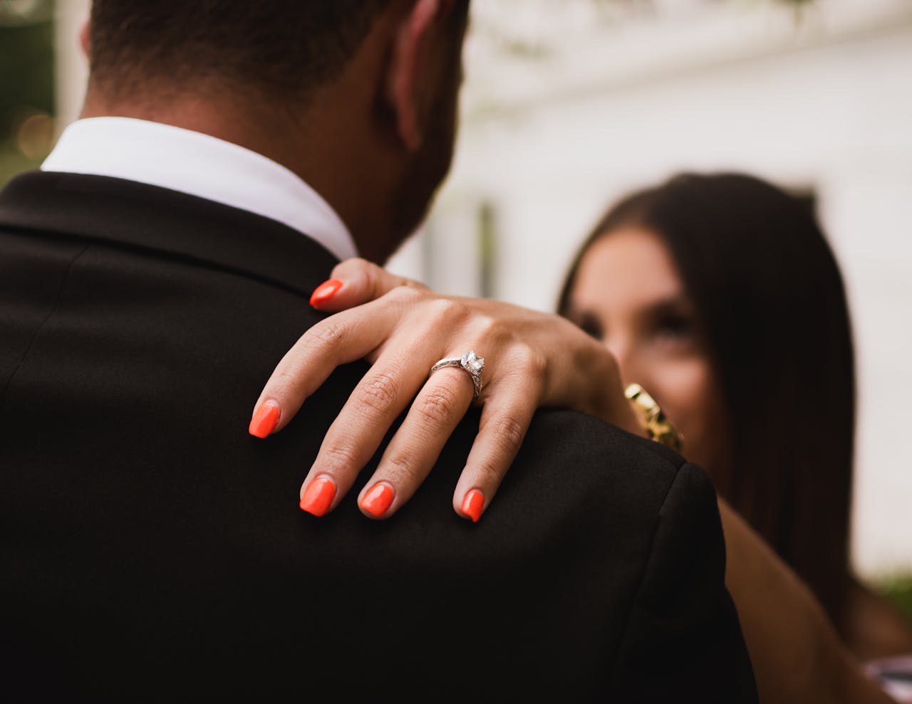 Close-up of a woman’s hand with a diamond ring resting on a man’s shoulder, both dressed formally outdoors.